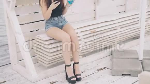 时尚女孩使用电话和杯状鸡尾酒在海滩长椅4K视频的预览图