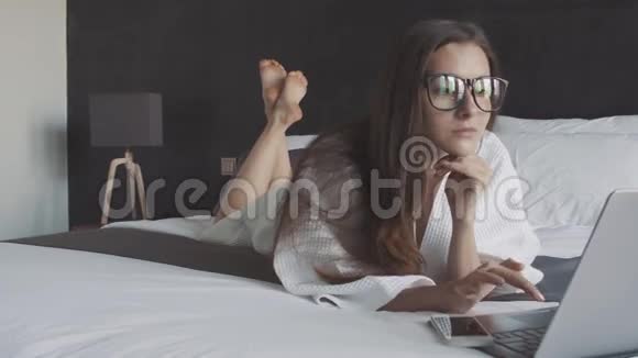 穿着白色浴袍和眼镜的女人在工作笔记本电脑躺在床上电脑上的智能手机在线商务视频的预览图