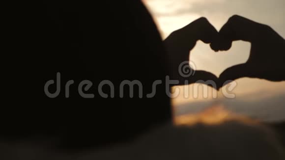 年轻美丽的女孩用她的手在心形框架夕阳日落时使心快乐的情感观念视频的预览图