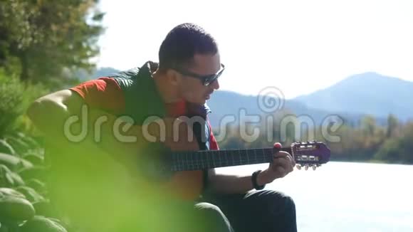 阳光明媚的一天戴着太阳镜的人坐在河边弹吉他唱歌3840x2160视频的预览图