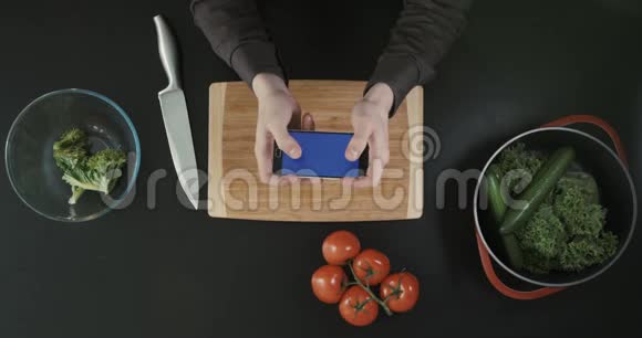 桌面上方手持手机的手的俯视图厨房概念砧板刀和蔬菜视频的预览图