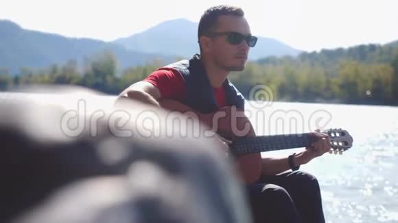 阳光明媚的一天戴着太阳镜的年轻人在坐在河边的吉他上缓慢地演奏3840x2160视频的预览图
