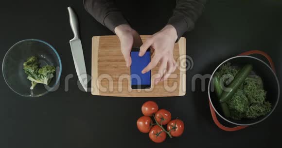上景手拿着蓝色屏幕的手机蔬菜砧板和刀在桌子上视频的预览图