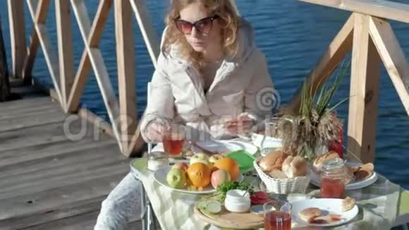年轻女子穿着暖和的衣服坐在桌子旁喝着茶吃着带果酱的煎饼在河边野餐视频的预览图