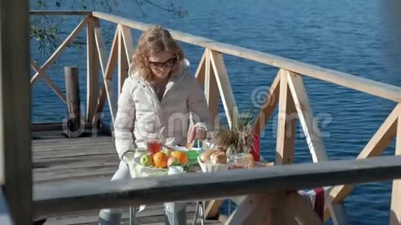 年轻女子穿着暖和的衣服坐在桌子旁喝着茶吃着带果酱的煎饼在河边野餐视频的预览图