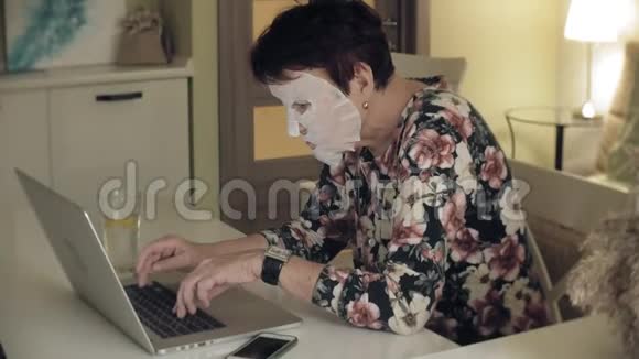 一位老妇人坐在桌子旁脸上涂了一张化妆纸巾她用电脑面部护理塑料视频的预览图