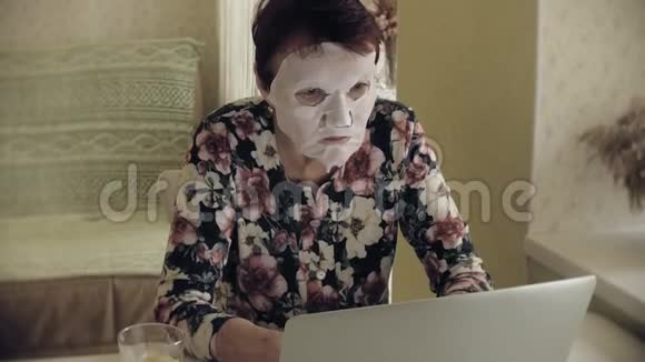 一位老妇人坐在桌子旁脸上涂了一张化妆纸巾她用电脑面部护理塑料视频的预览图