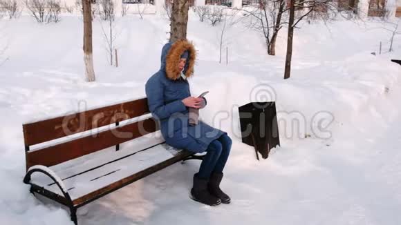 白天在下雪的天气里女人坐在长凳上在城市的冬季公园里浏览手机视频的预览图