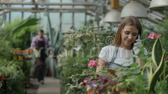 穿着围裙戴着手套的年轻开朗的女人一边打电话一边植物一边在温室里松开花视频的预览图