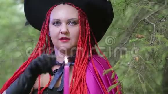 一个年轻的女巫红头发尖尖的帽子和黑色的斗篷在森林里用蜡烛变魔术视频的预览图