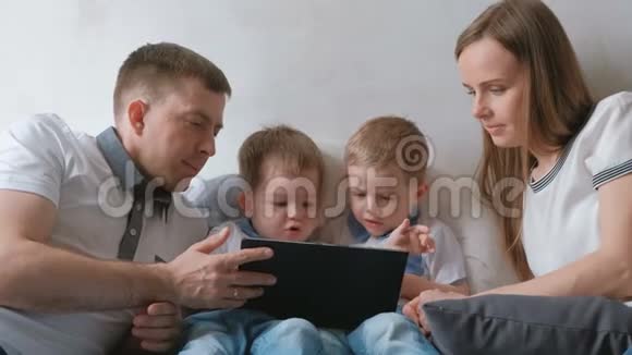带平板电脑的家人妈妈爸爸和两个儿子双胞胎蹒跚学步的孩子看着躺在床上的平板电脑视频的预览图