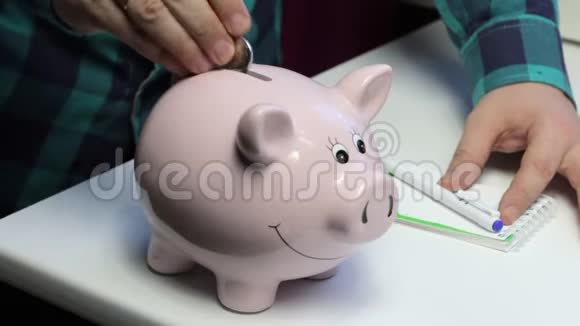 桌子上有一只粉红色的猪形的储蓄罐一个人把硬币放在里面在笔记本上做笔记视频的预览图