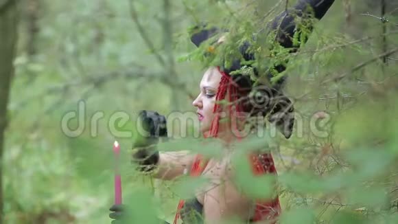 带着红色头发和尖尖帽子的迷人年轻女巫在森林里用蜡烛召唤视频的预览图