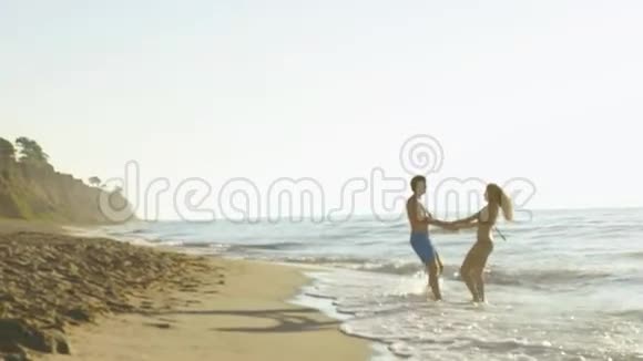 快乐的年轻夫妇在海边玩海浪在沙滩上英俊的男人和漂亮的女孩混在一起慢动作视频的预览图