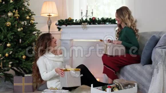 两个朋友打包好礼物坐在客厅里的女孩正在圣诞树附近聊天笑着视频的预览图