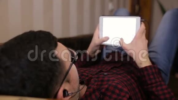 年轻人躺在沙发上在平板电脑上获取短信听音乐视频的预览图