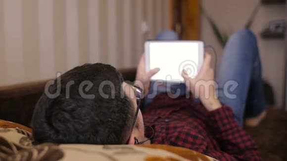 年轻人躺在沙发上在平板电脑上获取短信听音乐视频的预览图