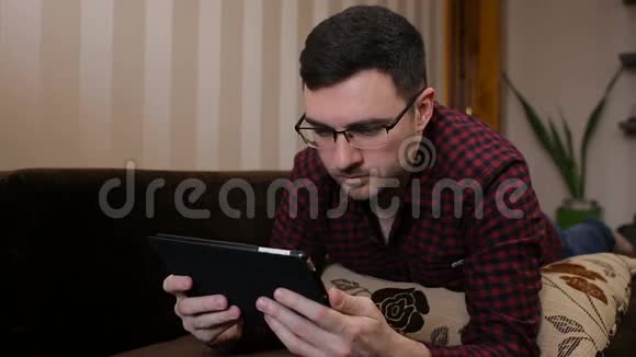 躺在沙发上有平板电脑的年轻人读着有趣的新闻视频的预览图