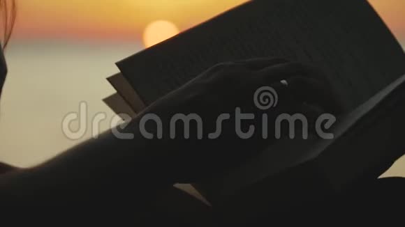 一个年轻女人坐在河边在日落时看一本书的剪影特写视频的预览图