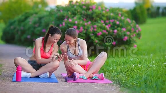 年轻活跃的健身女性在户外做运动看智能手机两个运动女孩在叙述中做运动视频的预览图
