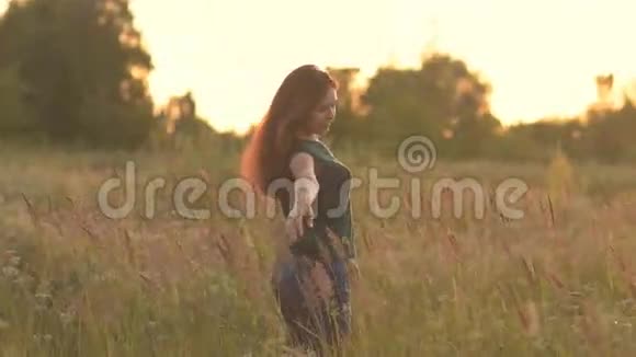 阳光下美丽的模特女孩穿着牛仔裤和绿色衬衫在田野上抚摸她的长发看着视频的预览图