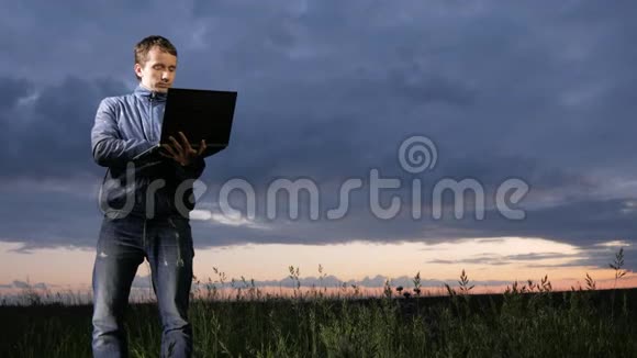 可爱的年轻人在田野日落时在笔记本电脑上工作背景上美丽的天空视频的预览图