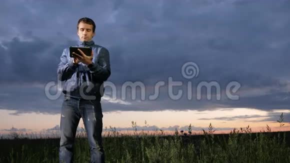 可爱的年轻人在田野日落时在平板电脑上工作背景上美丽的天空视频的预览图