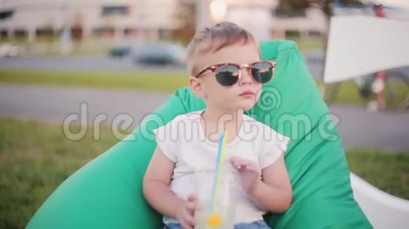 夏天可爱的小男孩戴着一副有趣的大太阳镜坐在公园的豆袋椅子上用吸管拿着杯子视频的预览图