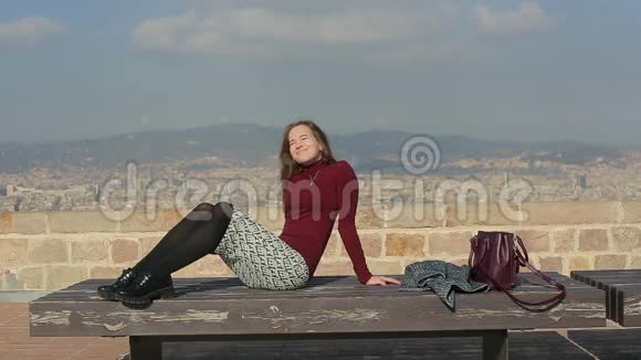 环顾四周微笑的女孩在城市顶端的长凳上晒太阳视频的预览图