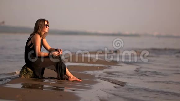 穿着黑色裙子和太阳镜的女孩坐在海滩上的岩石上向水里扔一颗鹅卵石视频的预览图