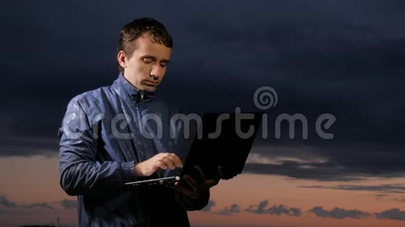 可爱的年轻人在田野日落时在笔记本电脑上工作背景上美丽的天空视频的预览图