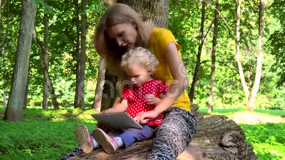 母亲带着蹒跚学步的女儿女孩用平板电脑坐在倒下的树干上视频的预览图