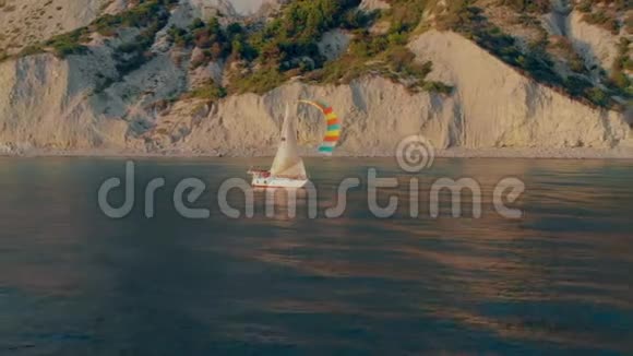 一艘白色的游艇在白色的帆下船员们平静地沿着蓝色的海洋沿着一个多山的海岸航行视频的预览图