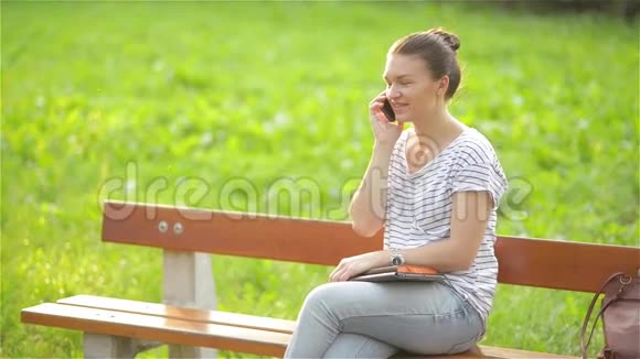 年轻漂亮的女人坐在城市公园的长凳上打着电话笑着说视频的预览图
