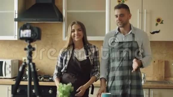 穿围裙的年轻有魅力的情侣在厨房里用dslr摄像机拍摄有关烹饪的视频食物博客视频的预览图