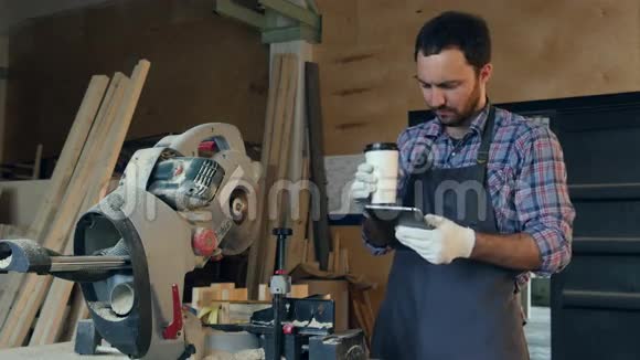 木匠在电锯附近的车间里喝咖啡和使用平板电脑视频的预览图
