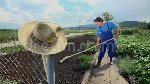 穿着蓝领衣服的人在花园里用耙子在地上移动视频的预览图