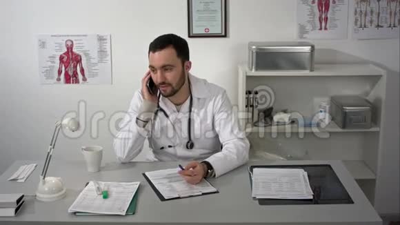 年轻的男医生坐在他咨询室的桌子旁用智能手机说话视频的预览图