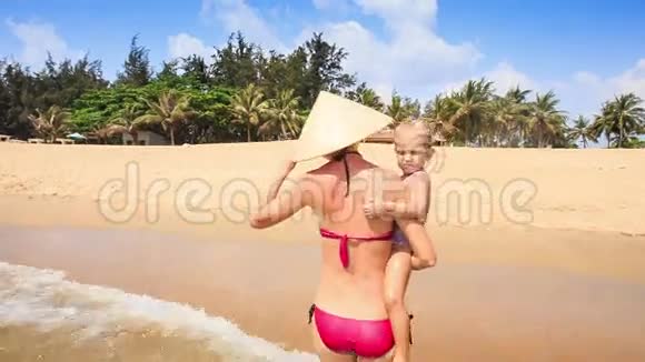 母亲在帽子里骑着手臂女儿沿着沙滩棕榈滩视频的预览图