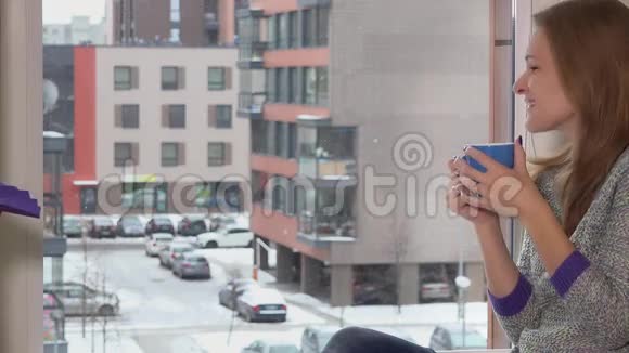 女人喝着一杯咖啡茶热饮坐在窗边回家下雪了视频的预览图