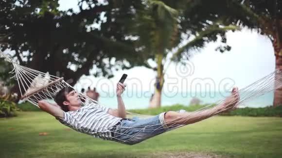 年轻男子在吊床上使用智能手机在美丽的热带海滩1920x1080视频的预览图