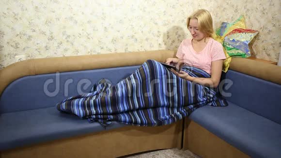 金发女人坐在沙发上用毯子盖着在平板电脑上玩视频的预览图
