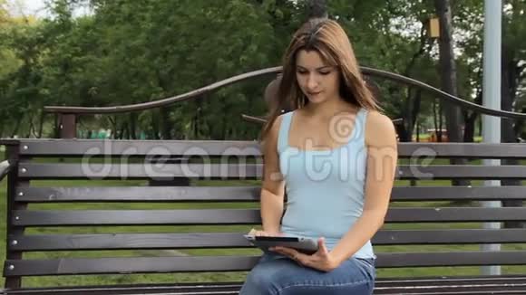 在城市公园用平板电脑的女人女人坐在公园里拿着小玩意的长凳上视频的预览图