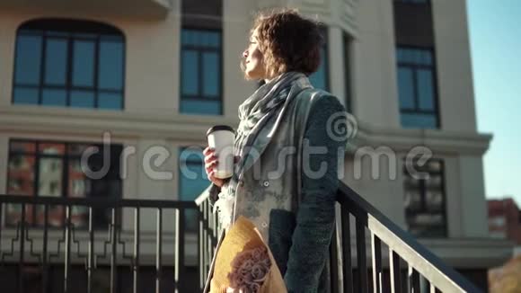 穿着城市背景外套的年轻女人带着一束鲜花的女孩喝咖啡视频的预览图