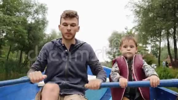 年轻的父亲和小儿子坐在湖边的船上年轻快乐的父亲和他可爱的孩子在木船上漂浮视频的预览图