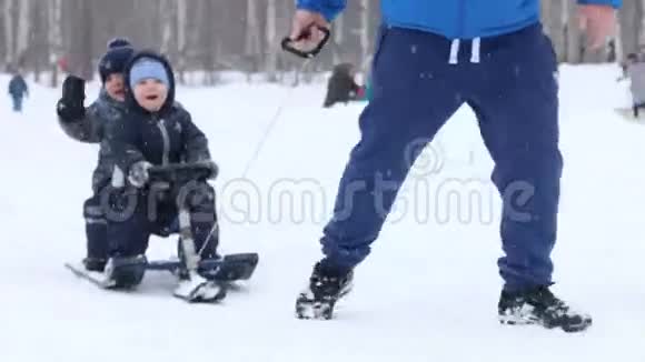 在公园下雪的时候雄性手拖着两个快乐的小男孩在雪橇上靠近其他男孩视频的预览图