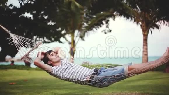 年轻的帅哥在美丽的热带海滩上的吊床上放松假期概念1920x1080视频的预览图