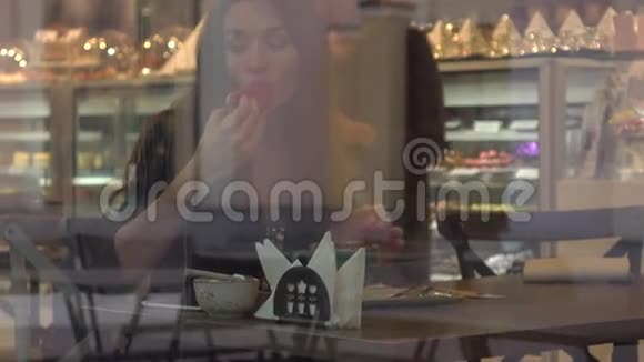 美丽的黑发年轻女子在糕点店吃蛋糕从外面透过窗户观看4K视频视频的预览图