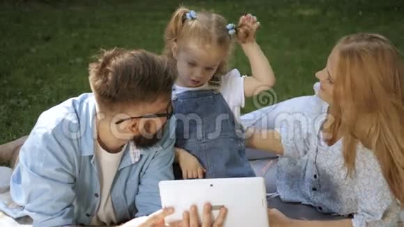 快乐女孩和爸爸妈妈在阳光明媚的夏日草地上玩耍的家庭画像和游戏平板电脑视频的预览图