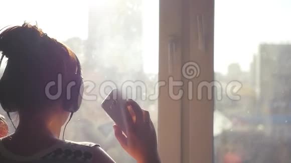 戴着大耳机的女孩在城市背景的窗户附近跳舞手里拿着手机的阳光1920x1080视频的预览图